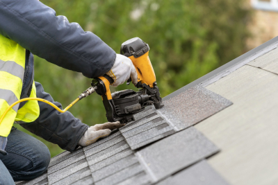 Person on roof wearing a utility vest with nail gun laying shingles.